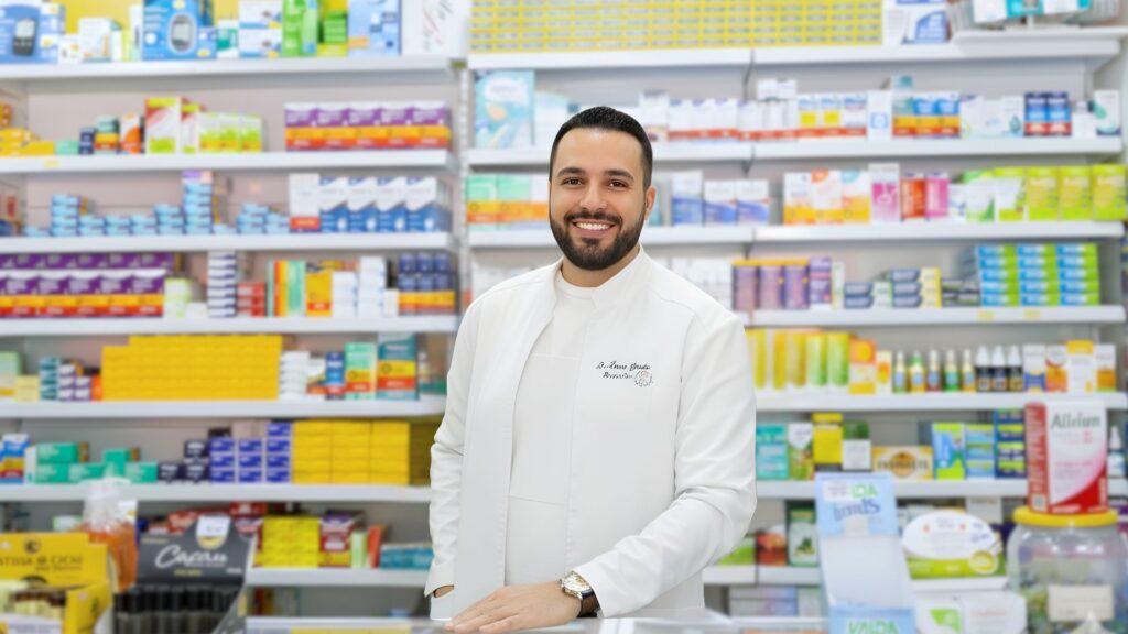 Farmacêutico sorridente da Farma e Farma atrás do balcão em farmácia moderna e organizada, simbolizando confiança e atendimento de qualidade.