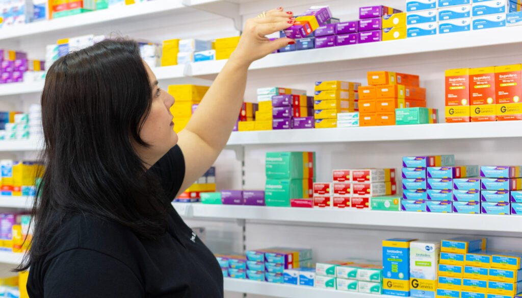 Mulher organizando medicamentos em prateleira de farmácia.